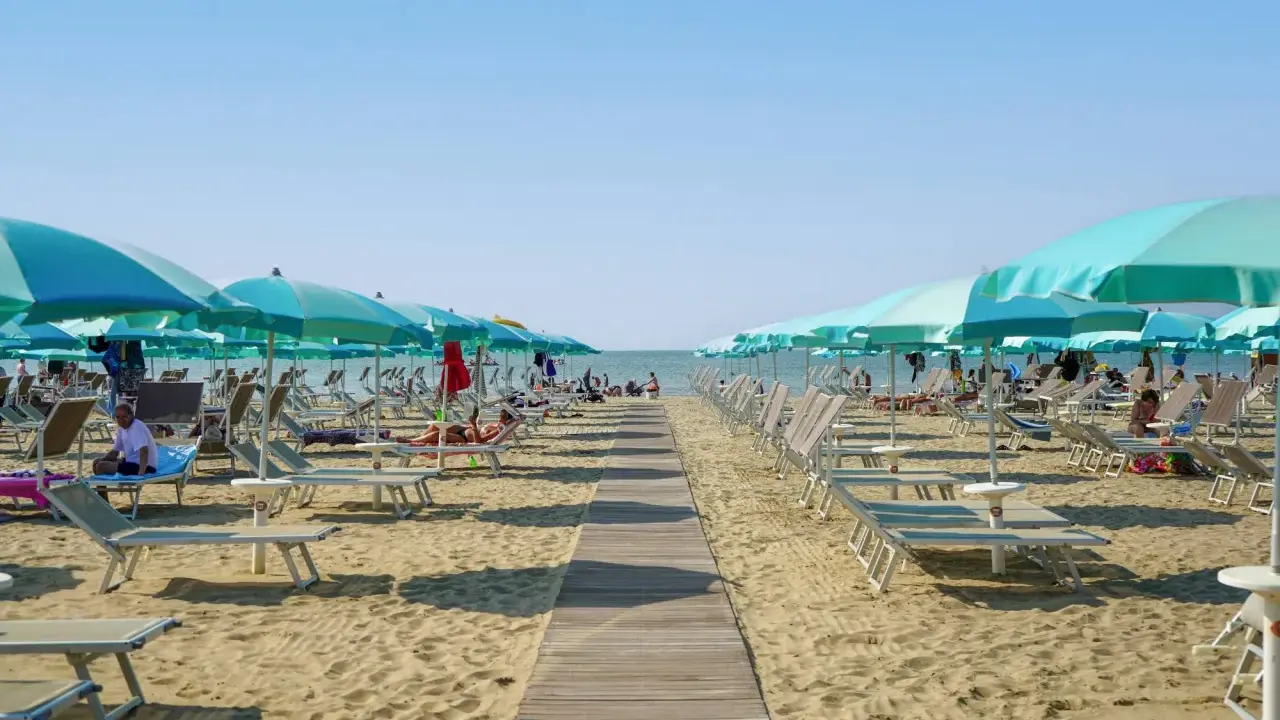 Una passerella di legno conduce alla spiaggia di San Giuliano Mare, fiancheggiata da file di lettini e ombrelloni azzurri sotto un cielo limpido.