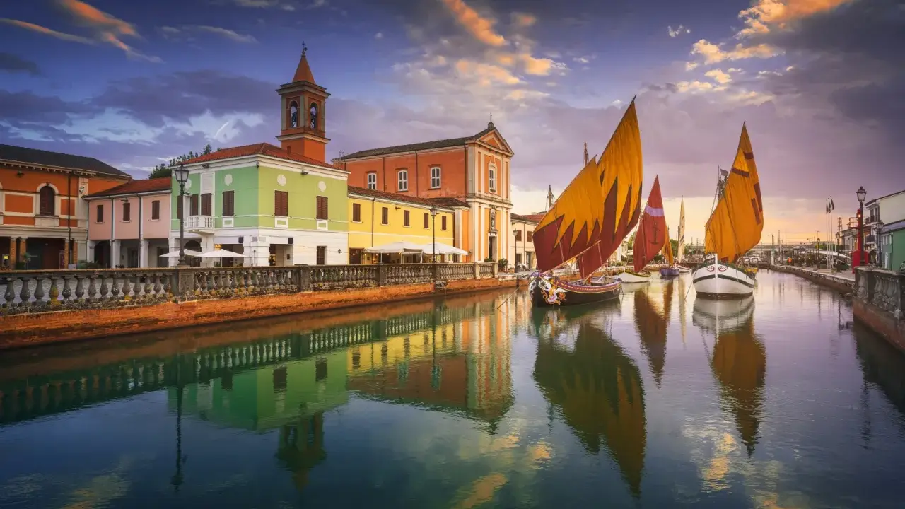 Navi a vela colorate navigano sul canale di Cesenatico al tramonto, con edifici vivaci e riflessi sull'acqua. Meglio Cervia o Cesenatico? Cesenatico offre questo spettacolo.