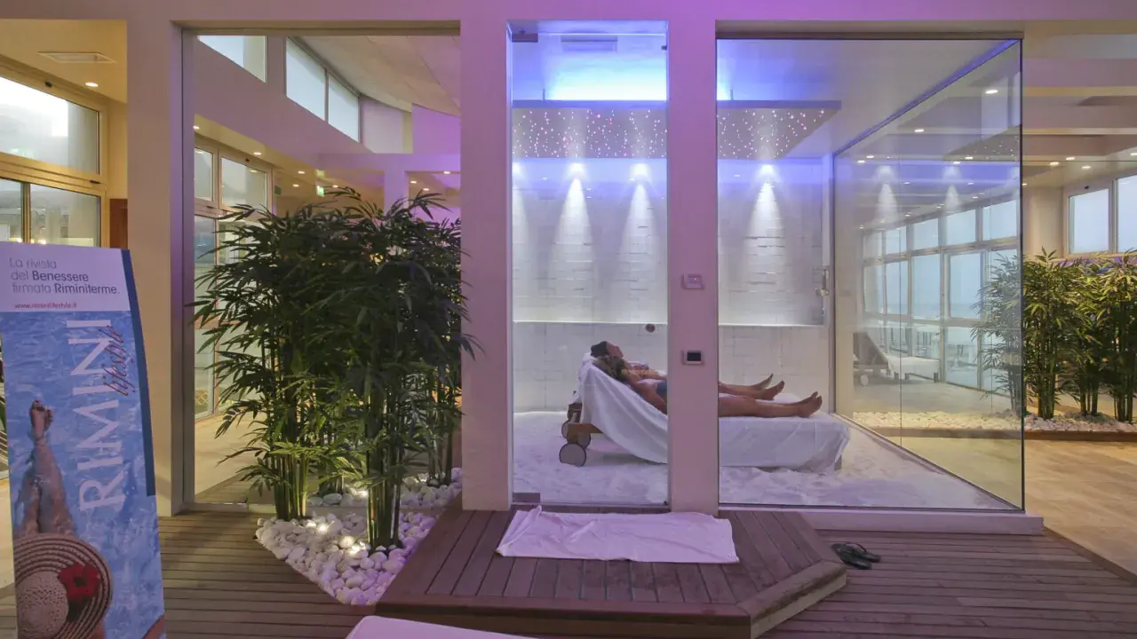 Un'oasi talassoterapico a Rimini, con due persone che si rilassano su lettini in un ambiente moderno e luminoso, circondato da piante di bambù e illuminazione soffusa.