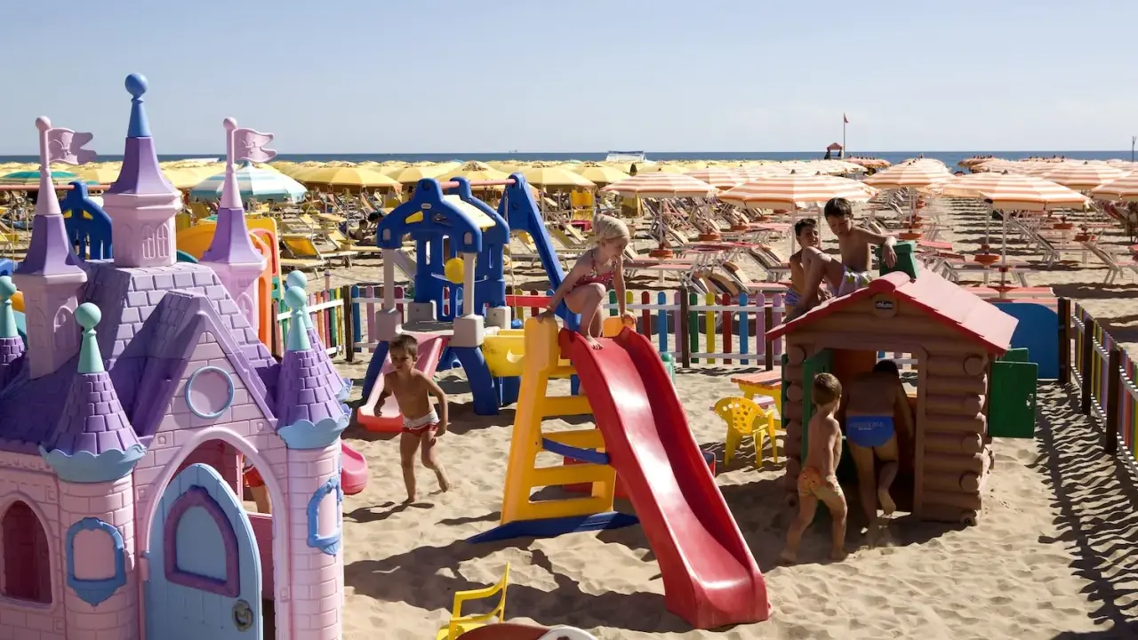 Bambini giocano in un'area giochi colorata sulla sabbia, con castello e scivoli, mentre ombrelloni gialli e lettini creano un'atmosfera da **sunday beach Rimini**.