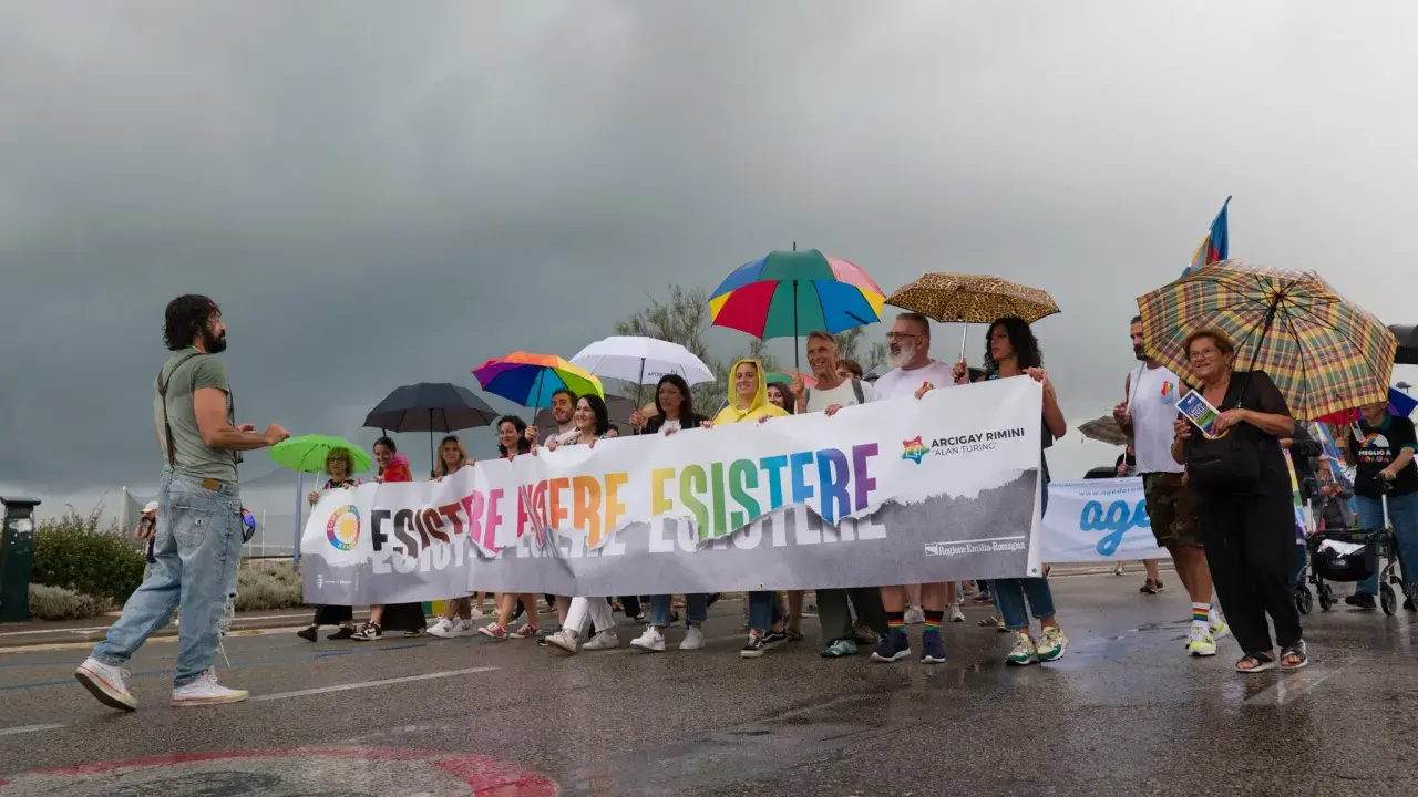 Persone con ombrelli colorati sfilano a Rimini Pride, tenendo uno striscione arcobaleno con scritto "Esistere Mere Esistere".