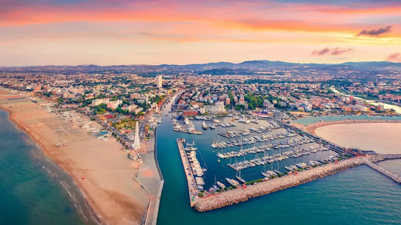 Tramonto sul lungomare di Rimini, con la spiaggia dorata, il porto turistico pieno di barche e la città che si estende fino alle colline.
