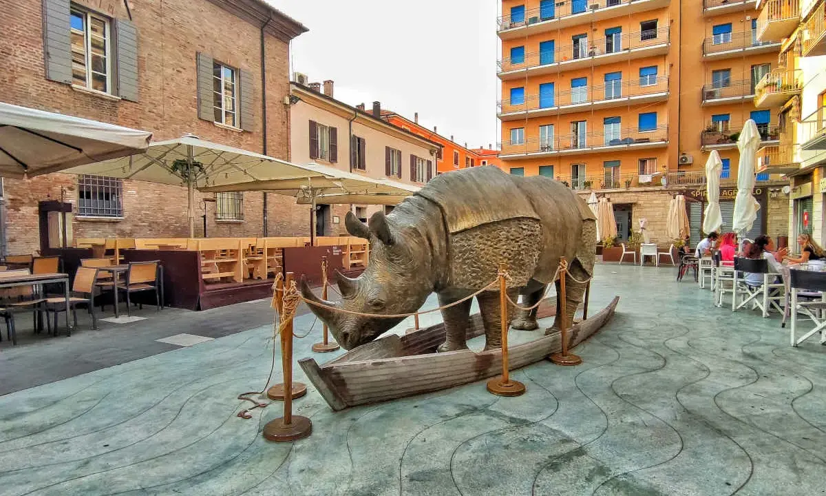 Statua di rinoceronte in una barca, in una piazza di Rimini, dove è nato Fellini. Tavolini all'aperto e palazzi colorati sullo sfondo.