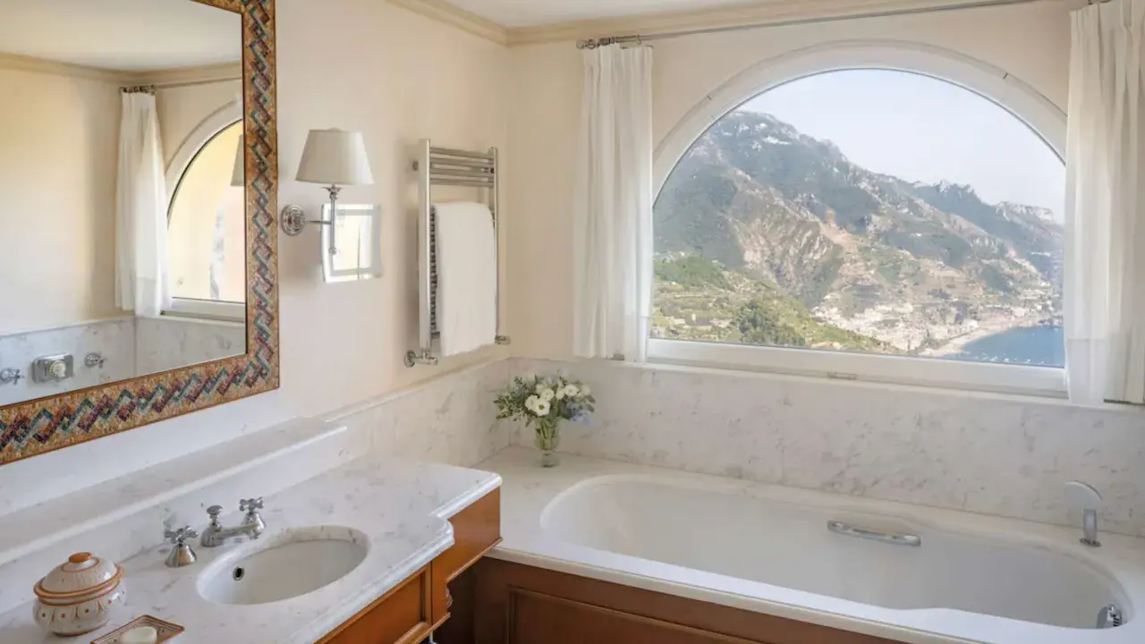 Un bagno di lusso con vasca, lavandino e specchio decorato, che offre una vista mozzafiato sulle montagne e sul mare. Tra i bagni più belli ed eleganti.