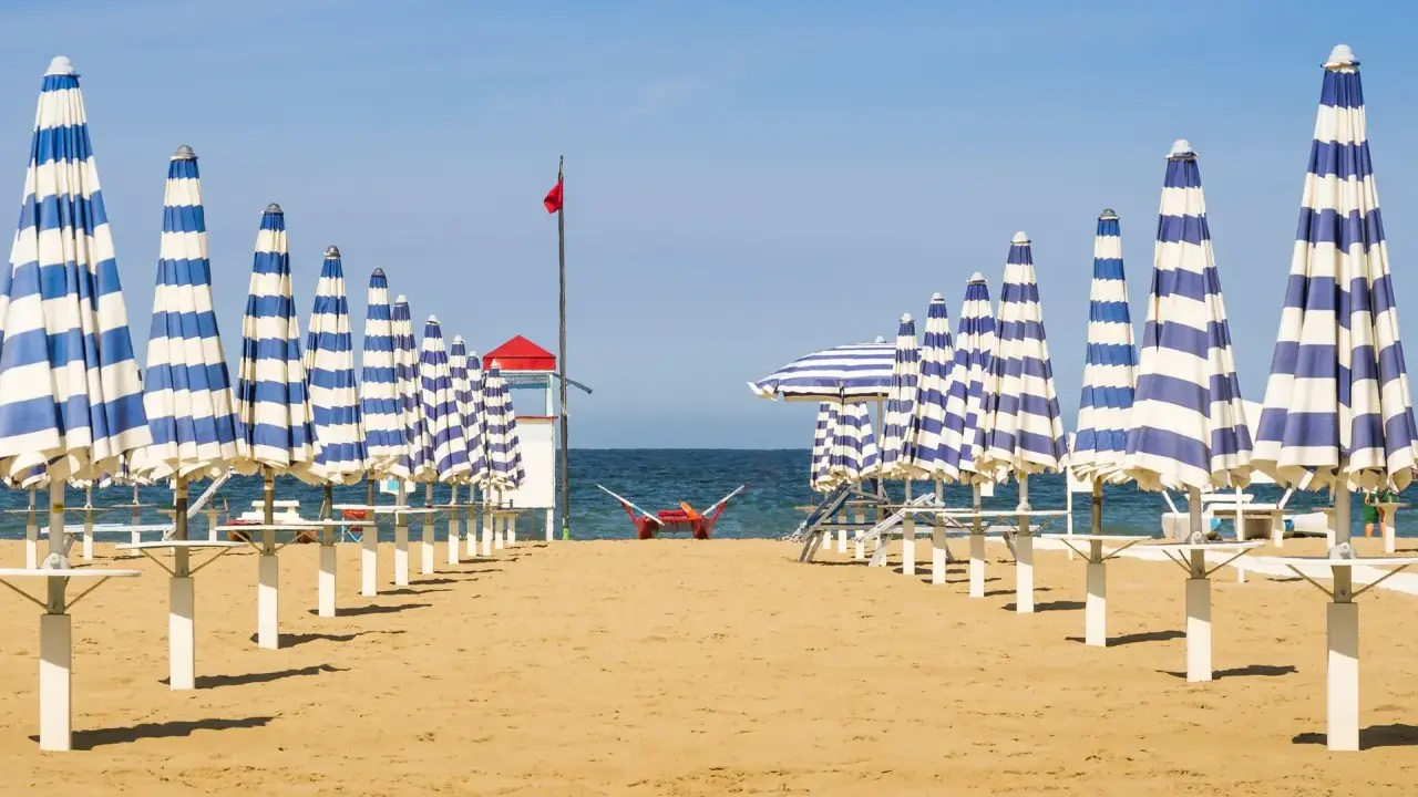 Filetto di ombrelloni a righe blu e bianche sulla spiaggia di Rivabella di Rimini, con mare calmo e cielo azzurro.