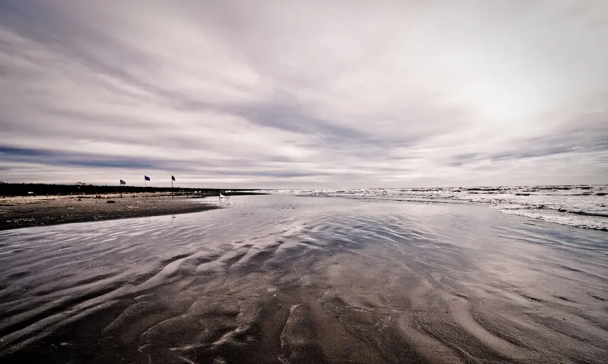 Spiaggia deserta con onde che lambiscono la riva, tre bandierine piantate nella sabbia bagnata. Un'atmosfera malinconica per questo mare d'inverno.