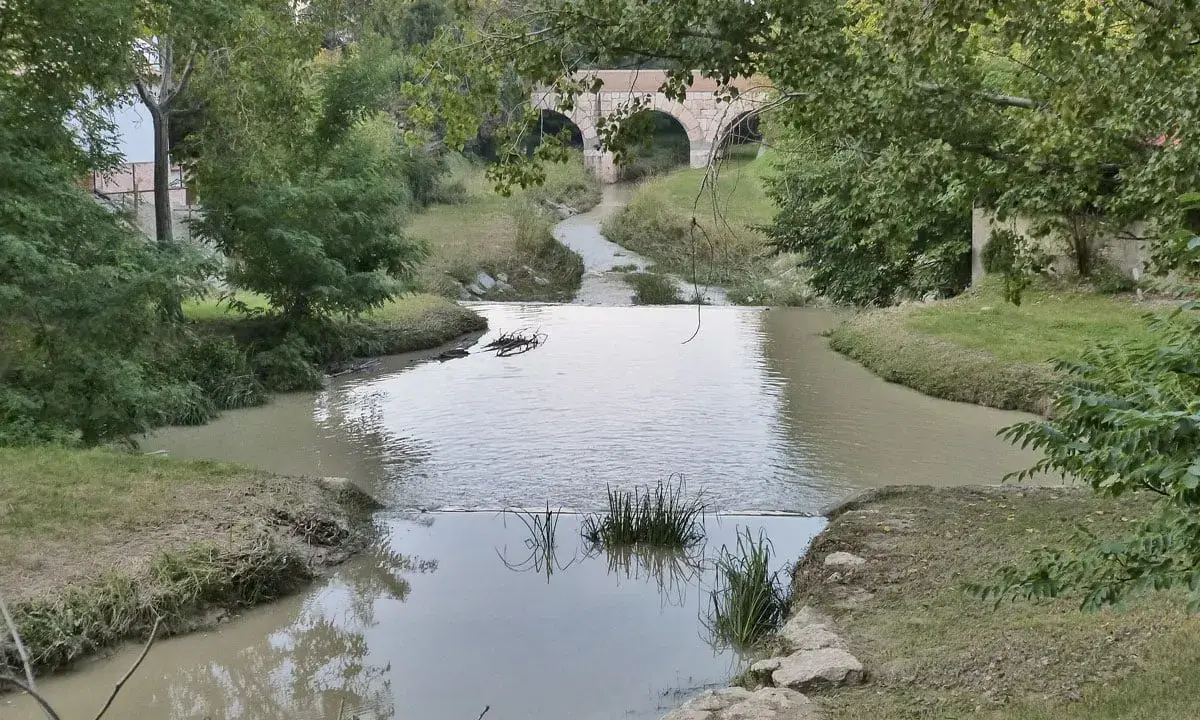 Un fiume serpeggia verso un antico ponte ad archi, evocando il **passaggio del Rubicone**. Vegetazione lussureggiante e acque tranquille.