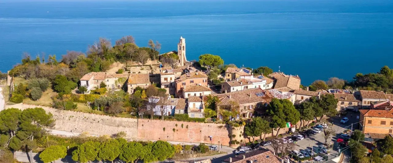 Borgo antico con campanile affacciato sul mare Adriatico. Il centro storico del lungomare Gabicce Mare, con tetti rossi e vegetazione rigogliosa, si estende verso l'azzurro intenso dell'acqua.