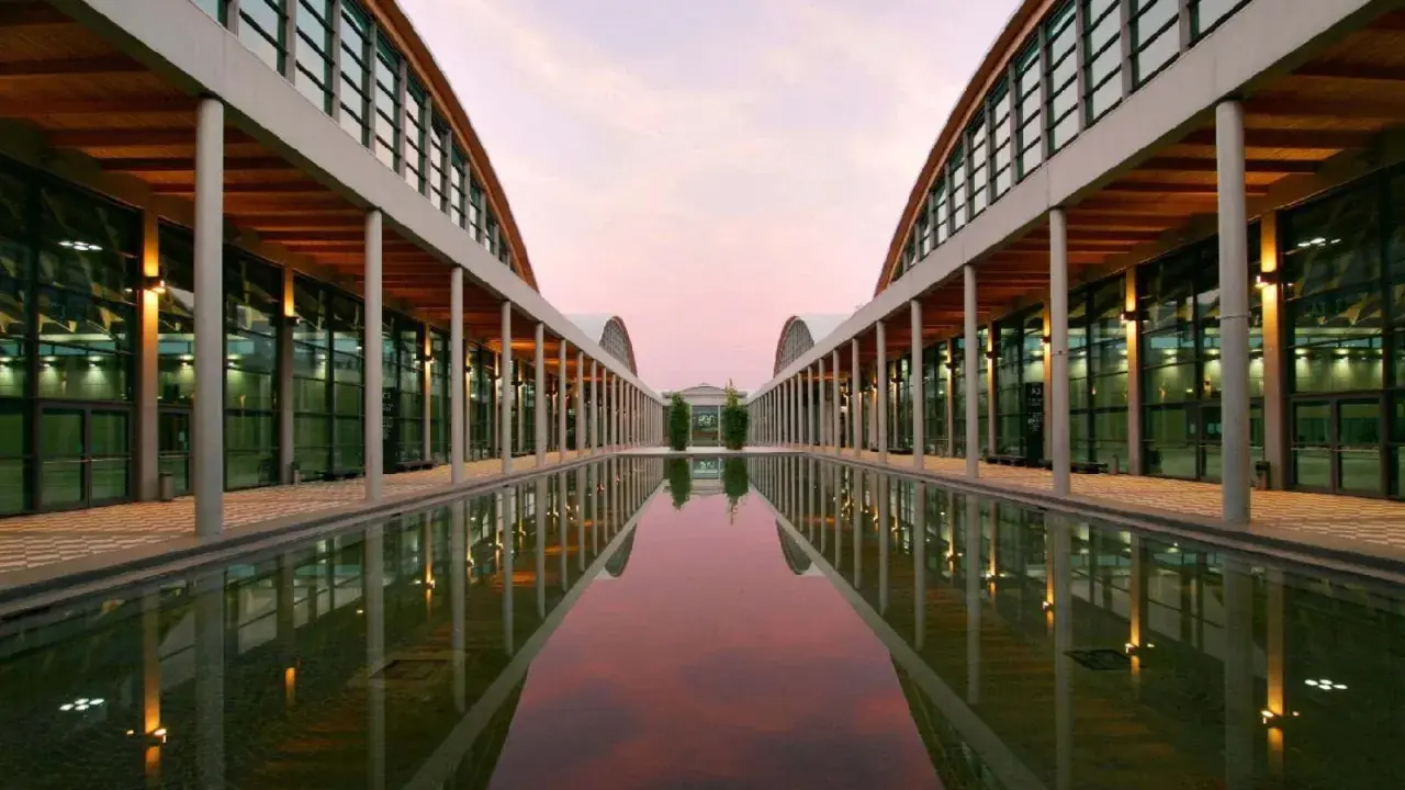 Ingresso moderno della fiera a Rimini, con un lungo specchio d'acqua che riflette le arcate e il cielo rosa del tramonto.