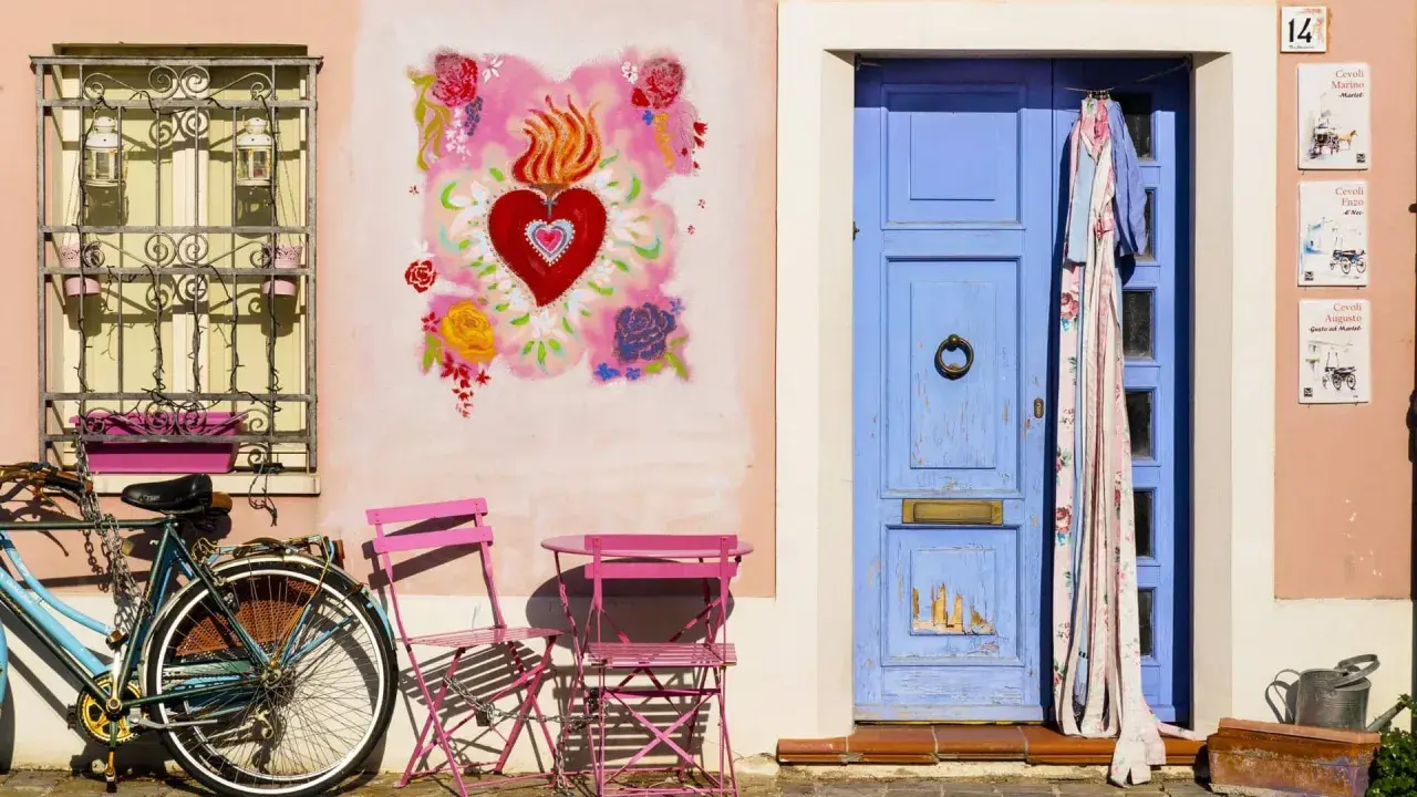 Un angolo pittoresco di Borgo San Giuliano a Rimini: una porta blu, un cuore fiammeggiante dipinto, una bicicletta e sedie rosa.
