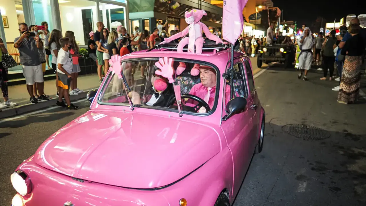 Un'auto rosa shocking sfila a Rimini durante la Notte Rosa, con un pupazzo a forma di pantera rosa sul tetto e un uomo al volante vestito a tema.
