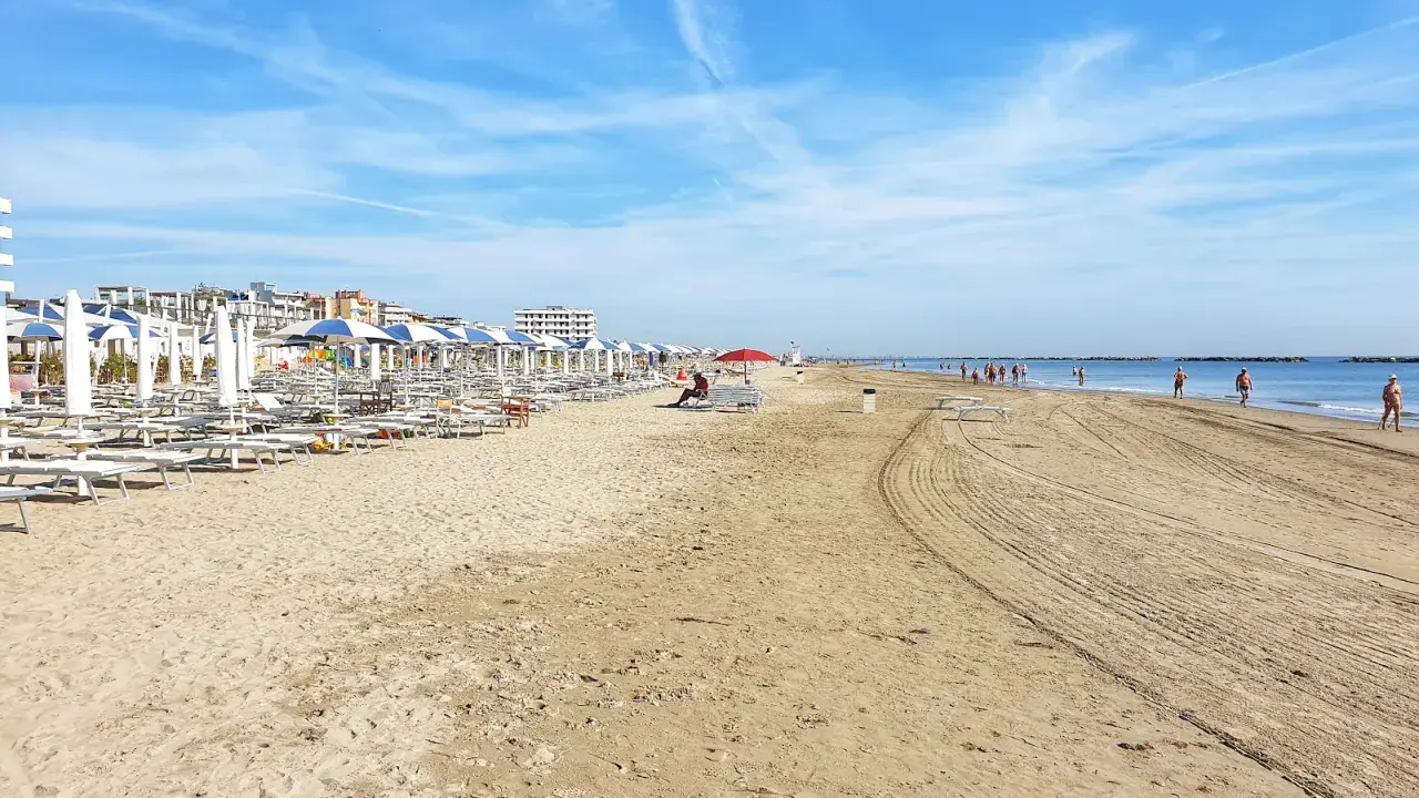 Spiaggia di Ravenna Lido Adriano con file di ombrelloni e lettini, bagnanti che passeggiano sul bagnasciuga sotto un cielo azzurro.