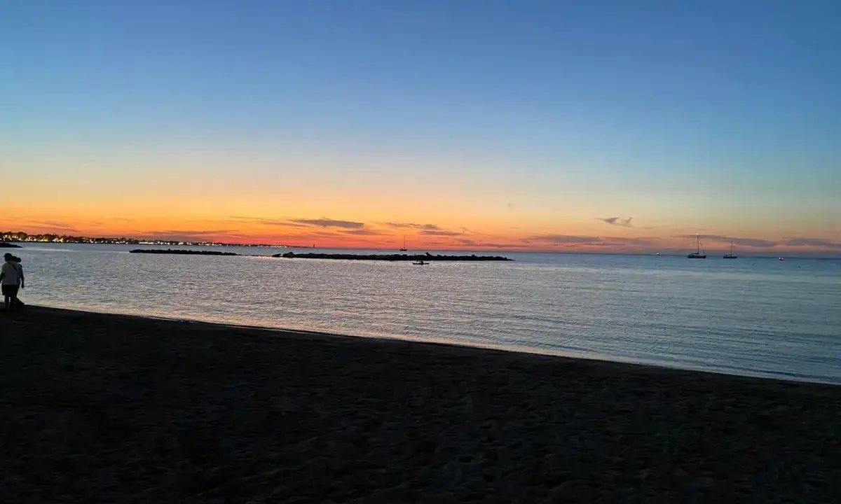 Tramonto sulla spiaggia di Rimini, con il cielo che sfuma dall'arancione al blu. Due persone passeggiano sulla sabbia scura, mentre in mare si vedono barche a vela.