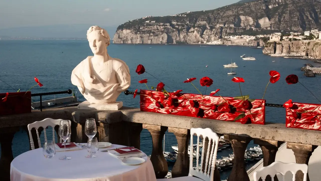 Tavolo apparecchiato con vista mare, perfetto per idee per Ferragosto. Busto in marmo, fiori rossi e panorama mozzafiato.