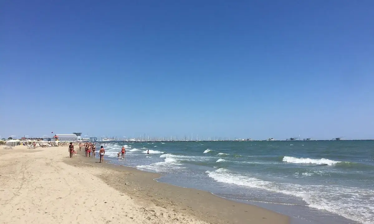 Spiaggia affollata a Ravenna, con gente che passeggia sulla sabbia e nel mare. All'orizzonte, barche e un cielo azzurro intenso.