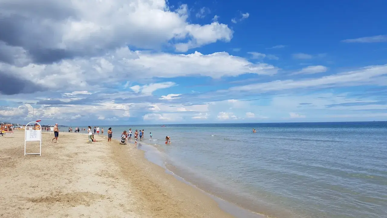 Una giornata di sole a Riccione mare, con gente che passeggia sulla sabbia e si gode l'acqua cristallina sotto un cielo azzurro con nuvole bianche.
