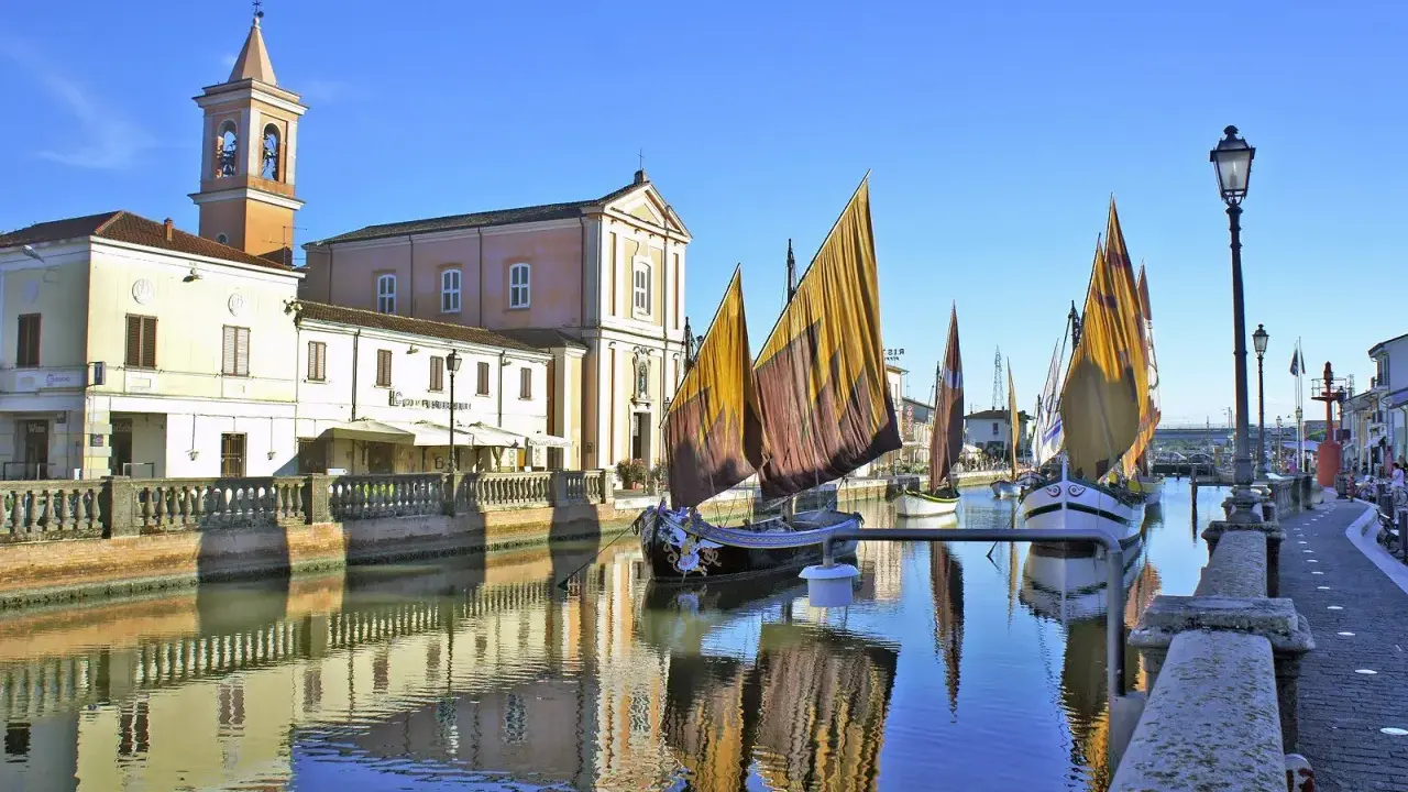 Traditional sailboats with colorful sails line a canal in Cesenatico, Italy, with buildings and a church reflected in the water.