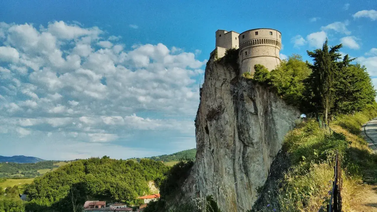 La fortezza di San Leo domina un picco roccioso, con un borgo sottostante e un cielo azzurro punteggiato di nuvole.