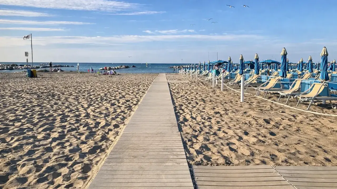 Passerella in legno sulla sabbia dorata di Rimini, con file di lettini e ombrelloni blu pronti per accogliere i bagnanti.