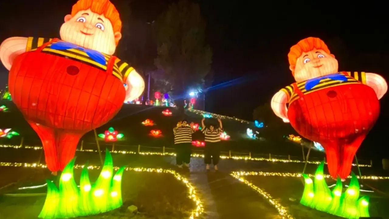 Gigantesche lanterne di bambini paffuti illuminano un sentiero a Wonderland Rimini, con due persone che salutano tra fiori luminosi.