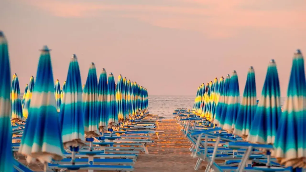 Filetto di ombrelloni a righe blu e bianche sulla spiaggia, con il mare e il cielo rosa all'orizzonte. Un'immagine iconica della riviera adriatica.