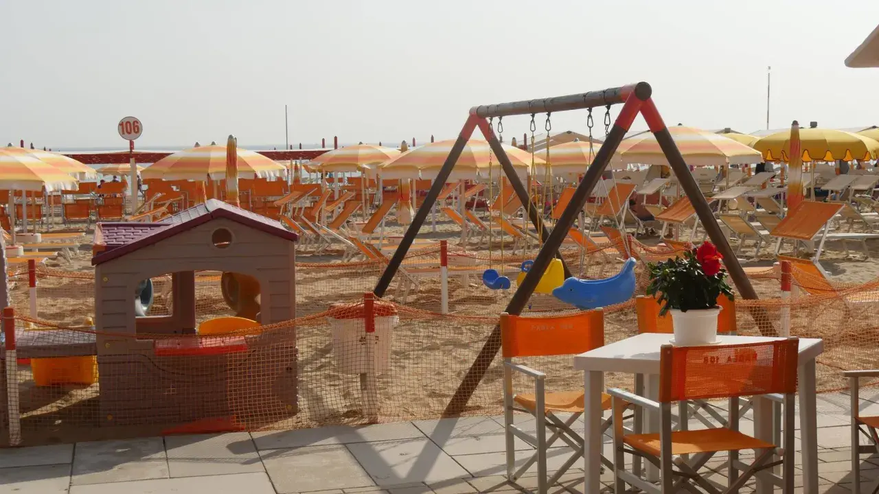 Area giochi e lettini sulla spiaggia 106 di Riccione, con ombrelloni arancioni e bianchi, altalena e casetta per bambini.