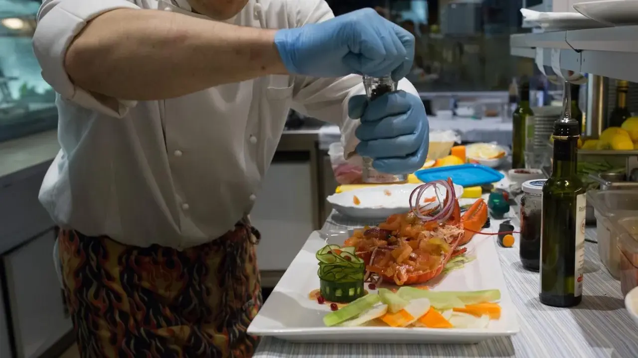 Chef con guanti blu macina pepe su un'aragosta, un piatto che sembra uscito da "lo zodiaco Rimini".