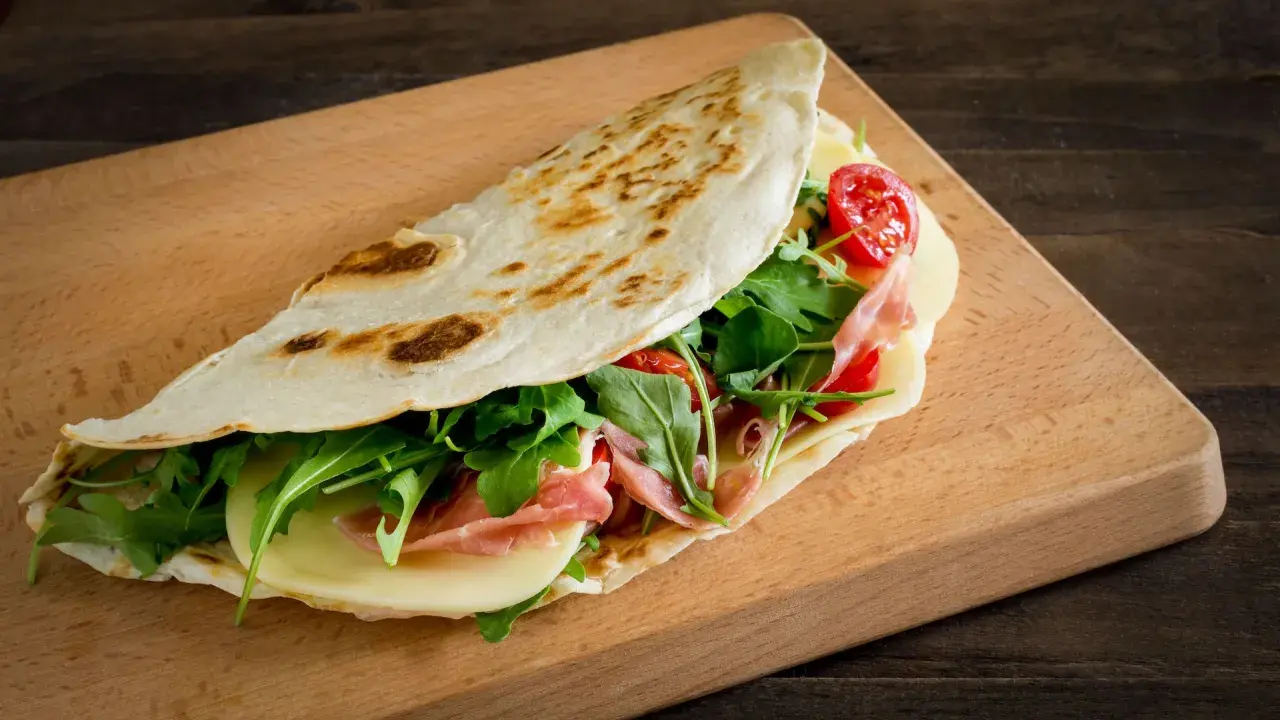 Piadina romagnola, un classico dal sapore autentico, ripiena di prosciutto, formaggio, rucola e pomodorini.
