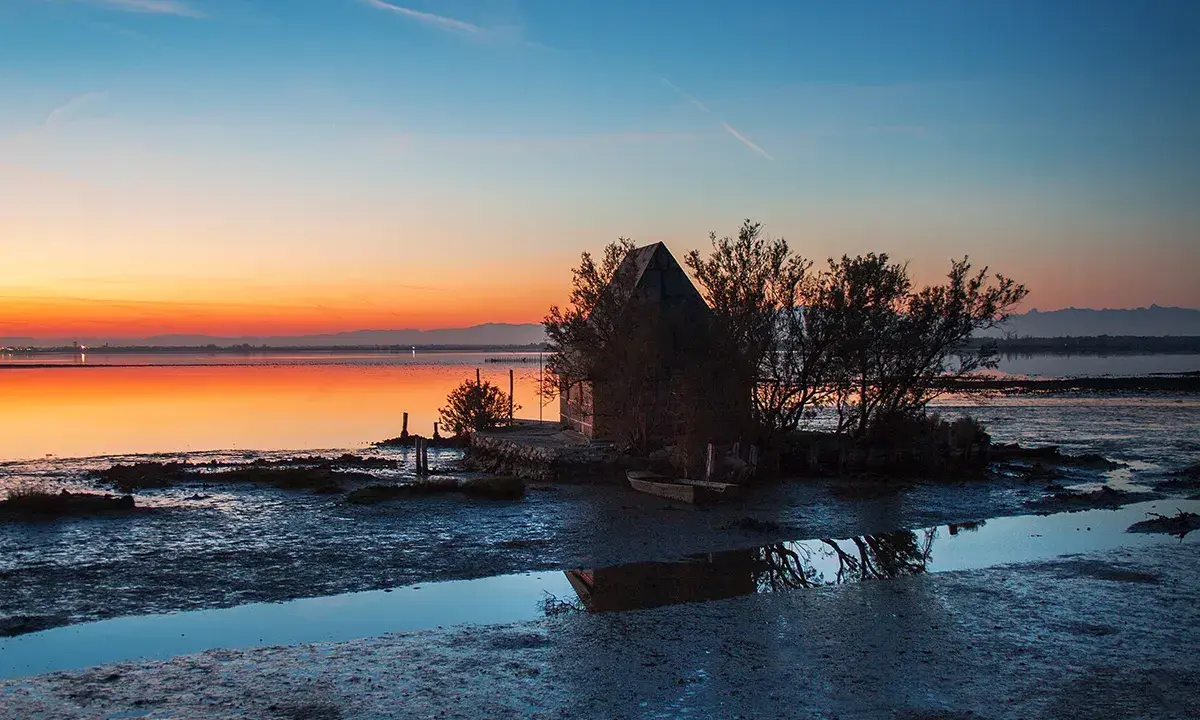 Tramonto sulla spiaggia di Marano, con una piccola capanna e un'imbarcazione riflessi sull'acqua calma.