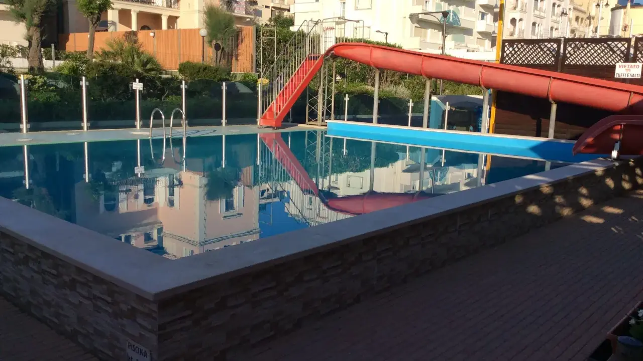 Piscina con scivolo rosso e giochi d'acqua ai Bagni Carmen Cattolica. Riflessi degli edifici nell'acqua azzurra.