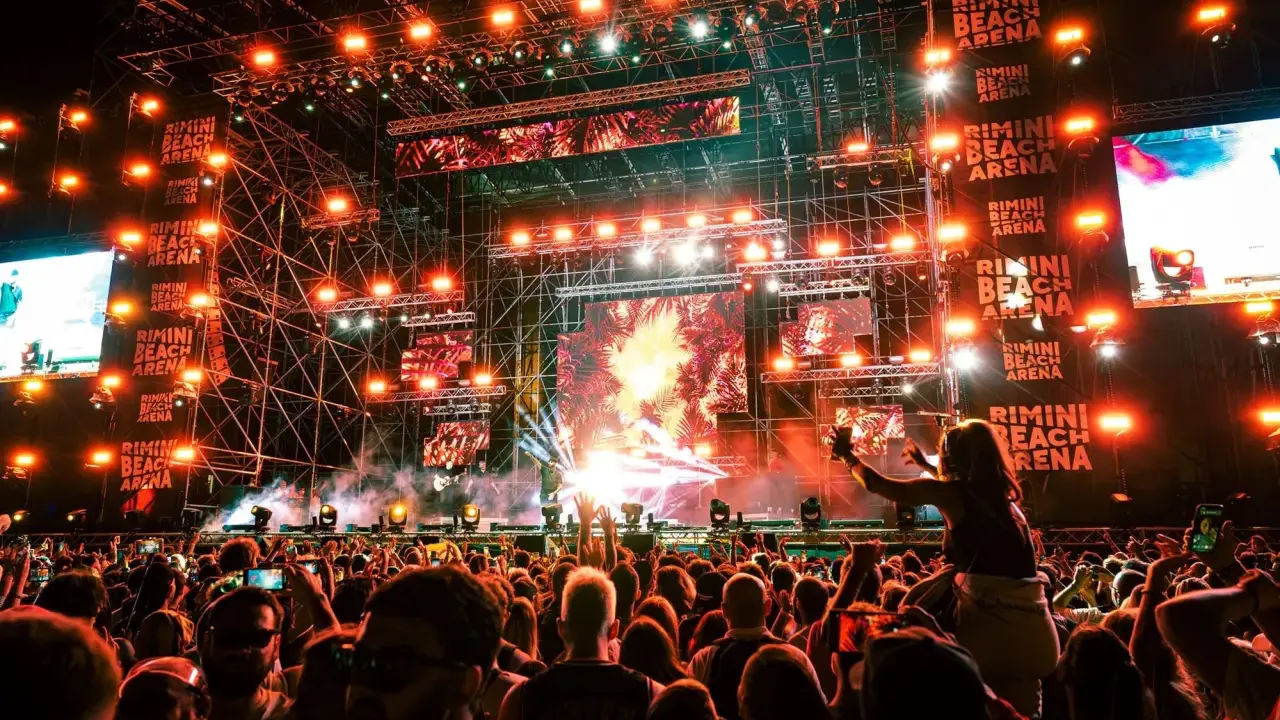 Folla in delirio al Rimini Beach Arena, luci psichedeliche e musica pulsante per un evento indimenticabile.