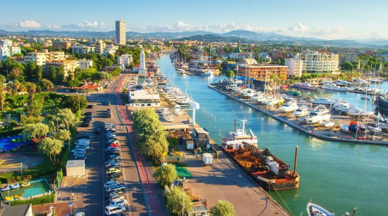 Vista aerea del porto di Rimini con barche ormeggiate, edifici e montagne sullo sfondo.