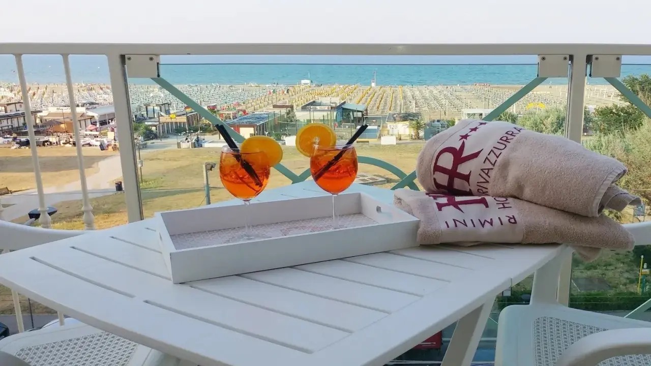 Due aperitivi arancioni e asciugamani con logo "Hotel Rivazzurra Rimini" sul tavolo, con vista sul rivazzurra mare e spiaggia attrezzata.
