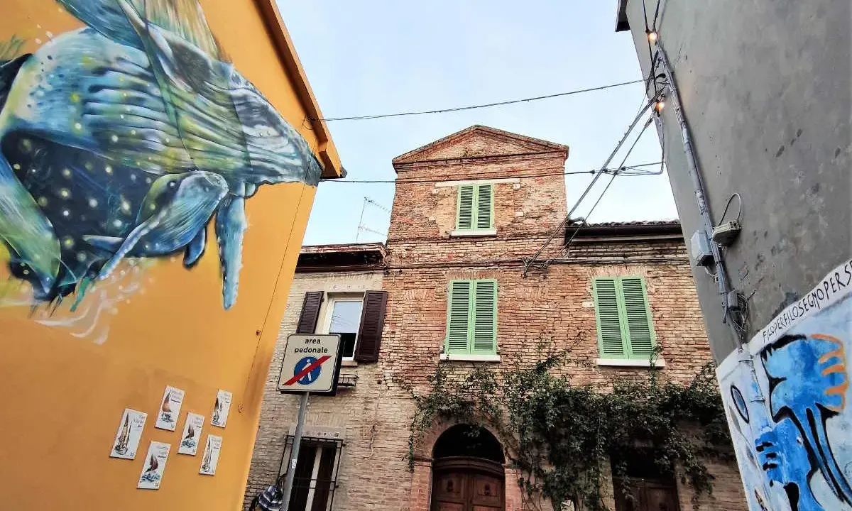 Un murale di balena colorata decora un edificio giallo a San Giuliano a Mare. Vicino, un edificio in mattoni con persiane verdi e un cartello di area pedonale.