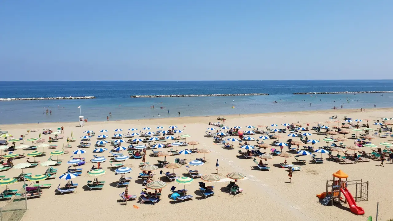 Spiaggia libera Viserba affollata di ombrelloni e lettini sotto un cielo azzurro. Bambini giocano nell'area attrezzata.