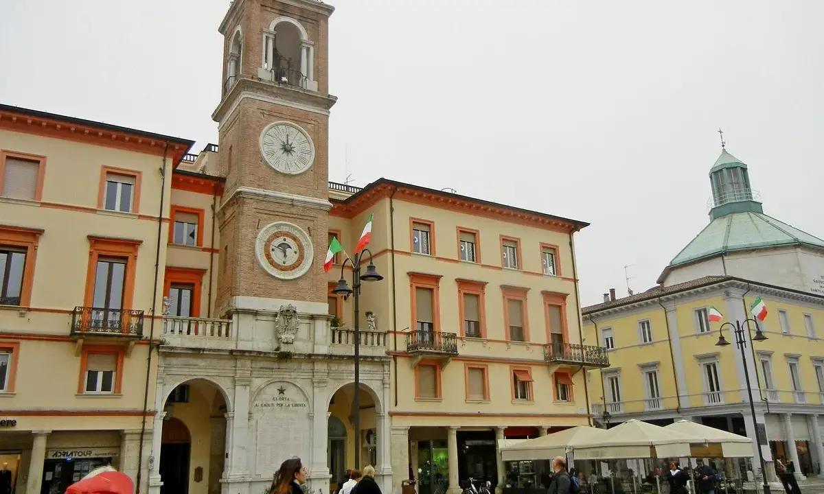 La piazza Rimini, con il suo imponente campanile e gli edifici storici, accoglie visitatori e residenti sotto un cielo nuvoloso.