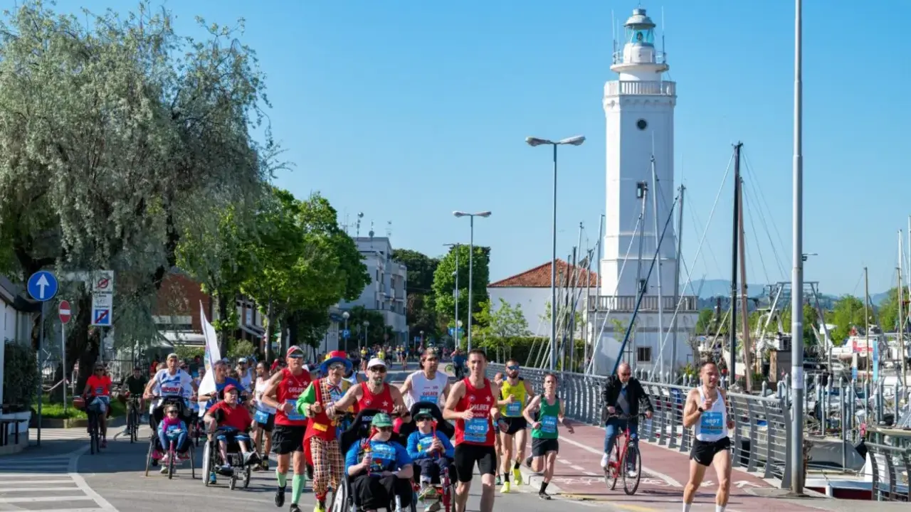 Atleti corrono lungo il percorso della Rimini Marathon, con il faro bianco e il porto sullo sfondo.