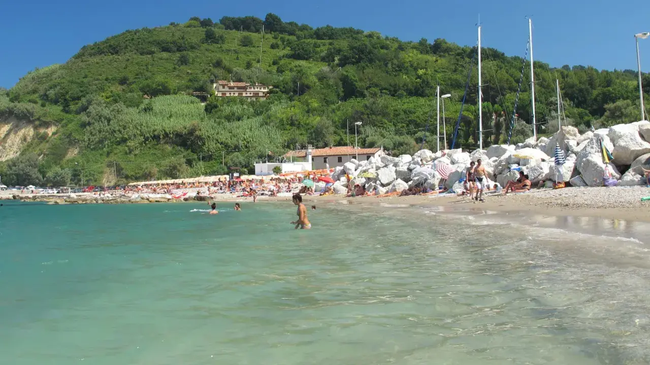 Spiaggia affollata con gente che nuota e prende il sole. Un promontorio verde domina la scena. Meglio Riccione o Gabicce Mare? Difficile scegliere!