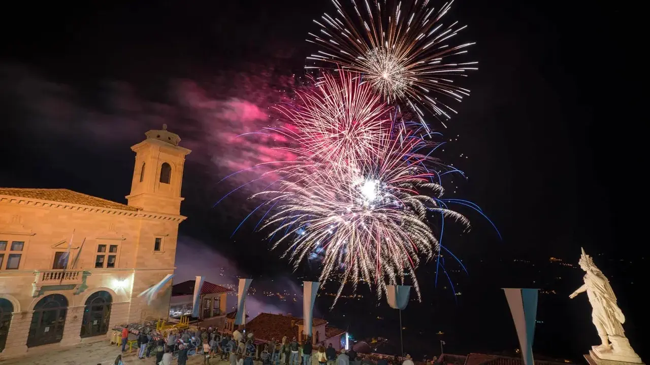 Spettacolo pirotecnico illumina il cielo notturno per la festa di San Marino. Folla ammira i fuochi d'artificio da una piazza con un edificio storico.