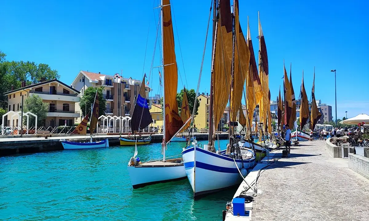 Barche a vela ormeggiate in un porto con case colorate sullo sfondo. Un dilemma: meglio Bellaria o Igea Marina?