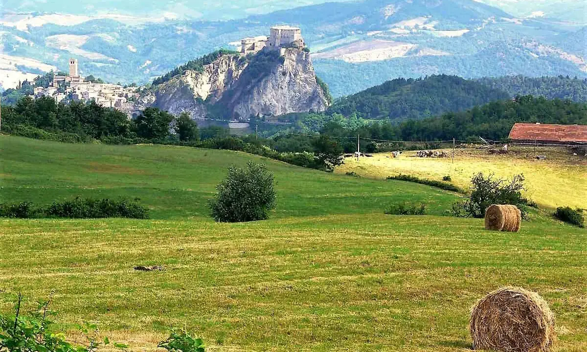 Campagna con borghi vicino Rimini, colline verdi, balle di fieno e un castello arroccato su uno sperone roccioso.