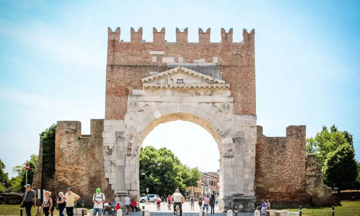 L'imponente arco d'augusto rimini, con le sue antiche pietre e la torre in mattoni, fa da sfondo a una vivace piazza con persone che passeggiano e ciclisti.