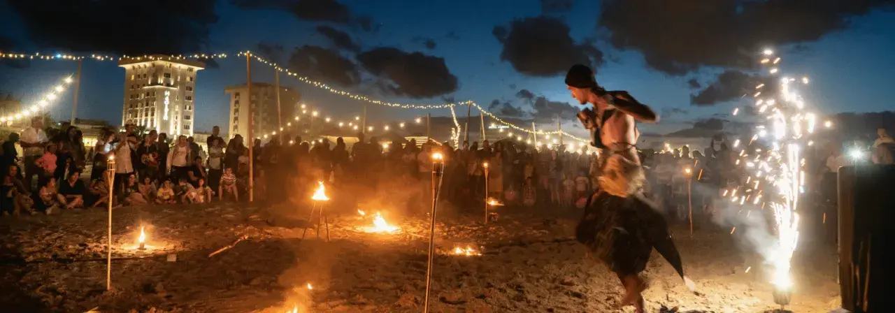 Spettacolo di fuoco sulla spiaggia di Rimini, estate. Artista si esibisce tra torce e fuochi d'artificio, con pubblico e luci in lontananza.