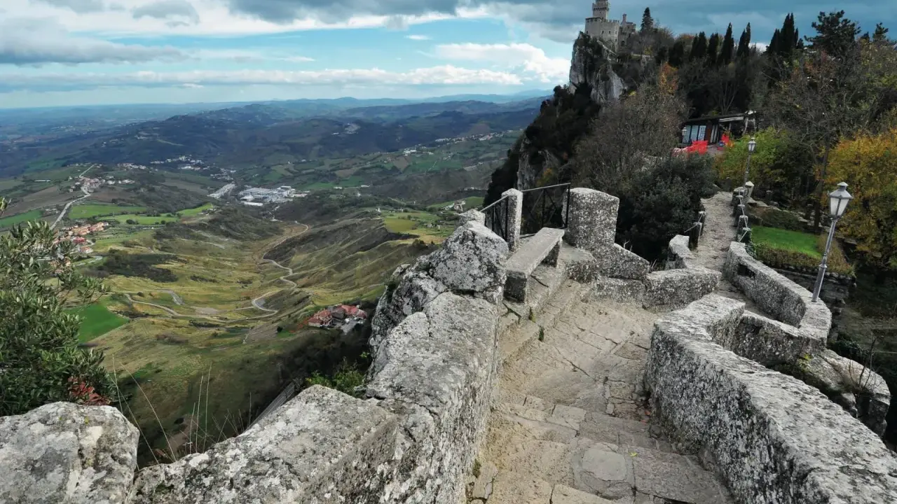 Sentiero in pietra conduce al **San Marino castle**, con vista panoramica sulla valle verdeggiante e colline ondulate sotto un cielo nuvoloso.