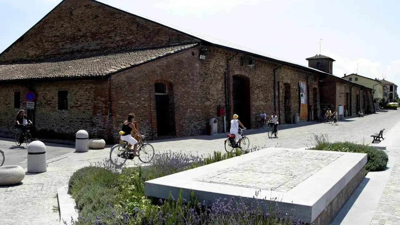 Passeggiata in bicicletta davanti ai Magazzini del Sale di Cervia, con persone che pedalano su un lungomare acciottolato, tra aiuole fiorite e antichi edifici in mattoni.