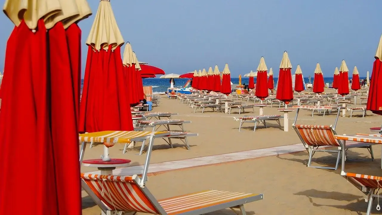 File di ombrelloni rossi e lettini prendisole su una spiaggia, forse a Riccione o Rimini, in attesa dei bagnanti.