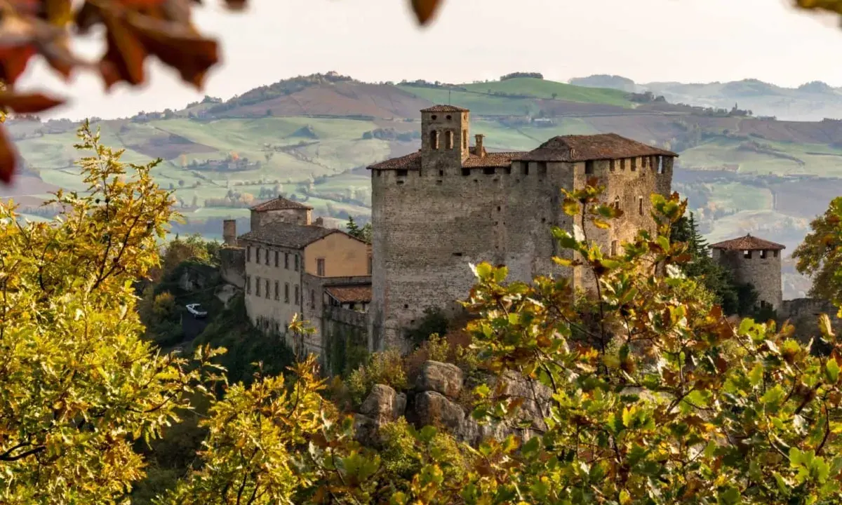 Castello medievale su una collina, circondato da alberi con foglie autunnali dai colori caldi. Il paesaggio collinare sullo sfondo evoca un senso di pace in questo mare autunno.