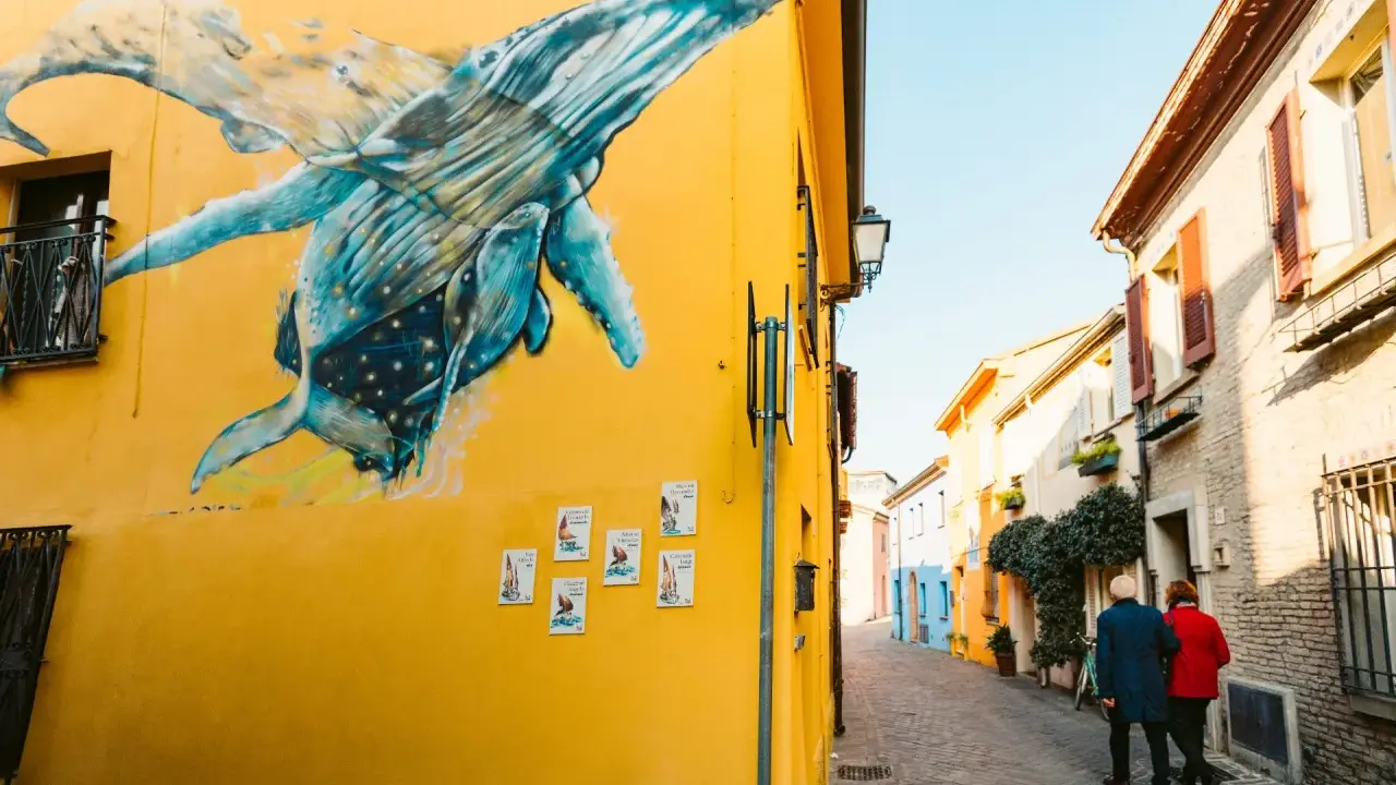 Un murale di balene blu su un muro giallo a San Giuliano Rimini. Una coppia passeggia lungo la strada acciottolata.