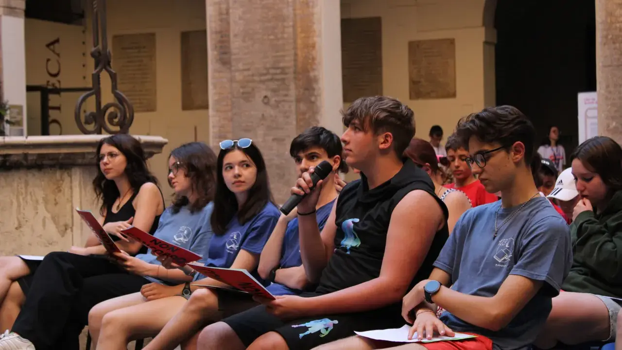 Giovani ascoltano un discorso, alcuni con fogli rossi in mano, durante un evento culturale a Rimini. Un ragazzo tiene un microfono.