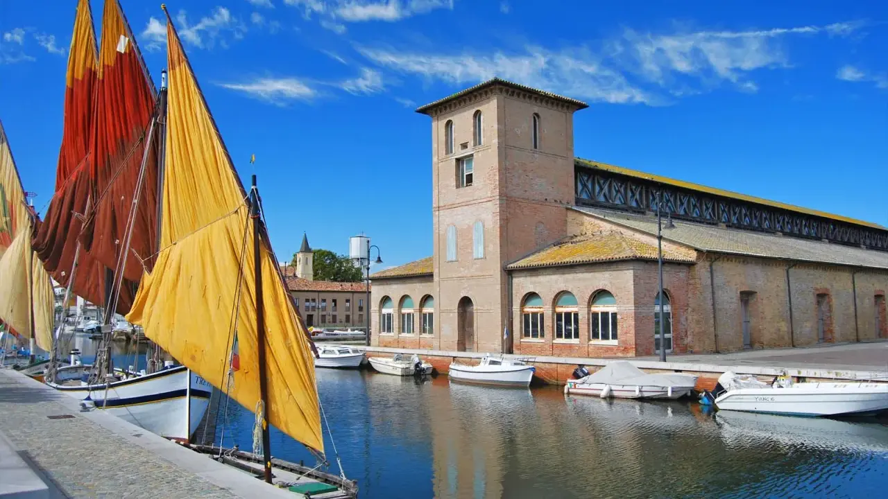 Vele colorate di barche tradizionali ormeggiate nel porto canale di Cervia mare, con l'antico edificio delle saline sullo sfondo.